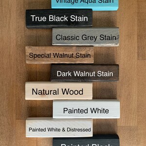 May include: Nine wooden blocks stacked on top of each other, each block has a different color and text. The colors are aqua, black, gray, brown, dark brown, natural wood, white, white with distressed paint, and black. The text on the blocks reads: Vintage Aqua Stain, True Black Stain, Classic Grey Stain, Special Walnut Stain, Dark Walnut Stain, Natural Wood, Painted White, Painted White & Distressed, Painted Black.