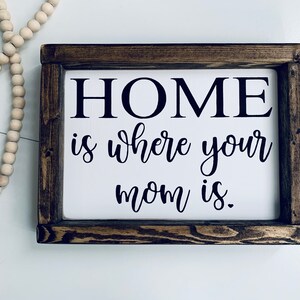 May include: A wooden sign with a white background and black text that reads "HOME is where your mom is."