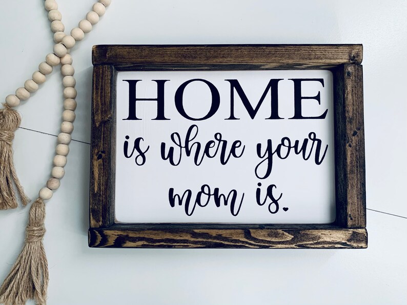 May include: A wooden sign with a white background and black text that reads "HOME is where your mom is."