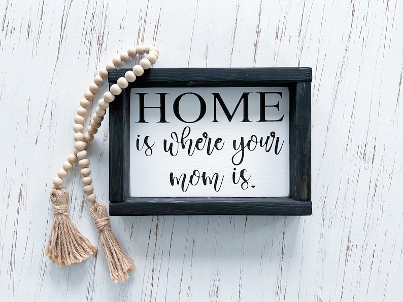 May include: A black framed sign with white background and the text "HOME is where your mom is." The sign is on a white wood background with a wooden bead garland with tassels.