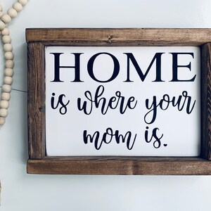 May include: A wooden sign with a white background and black text that reads "HOME is where your mom is."