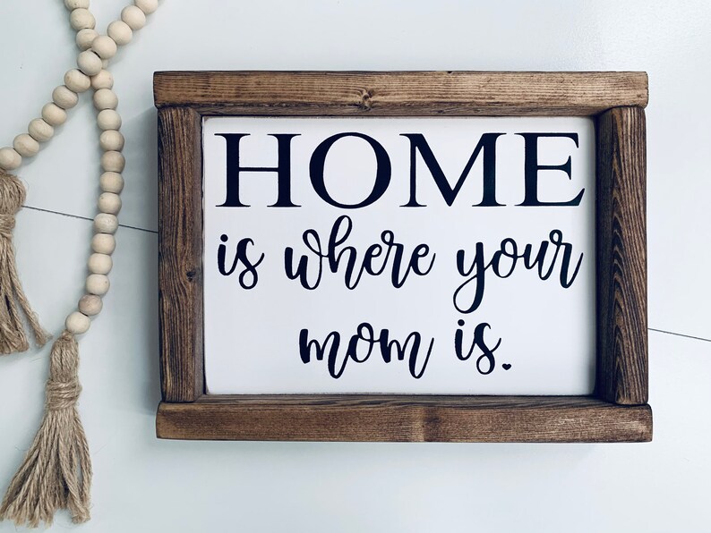 May include: A wooden sign with a white background and black text that reads "HOME is where your mom is."