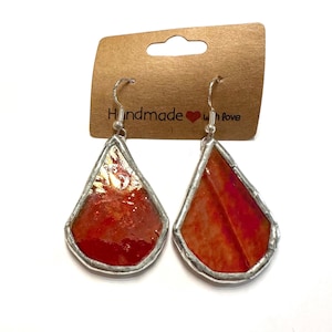 May include: A pair of handmade, teardrop-shaped earrings with a vibrant red and orange color scheme. The earrings have a silver-colored metal border and a hook closure. The backing card reads "Handmade with love."