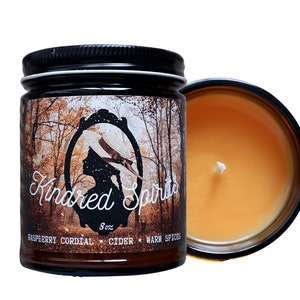 May include: A scented candle in a glass jar with a black lid. The label features a silhouette of a woman in a hat and the words "Kindred Spirits." The candle is 8 oz and has a warm, amber-colored wax. Scents include raspberry cordial, cider, and warm spices.