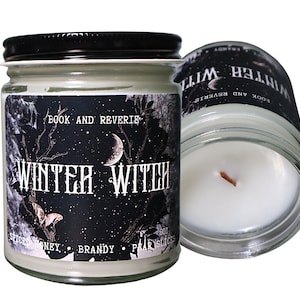 May include: Two glass jar candles with black lids. The labels read "Winter Witch" with "Book and Reverie" above and "Spiced Honey, Brandy, Pear Slices" below. One candle is upright, the other tilted.