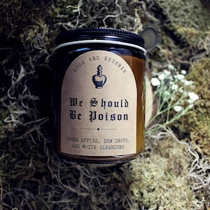 May include: A candle in a glass jar with a black lid, set against a backdrop of moss and greenery. The label reads "Book and Reverie" and "We Should Be Poison", with scent notes of green apples, dew drops, and white oleanders.