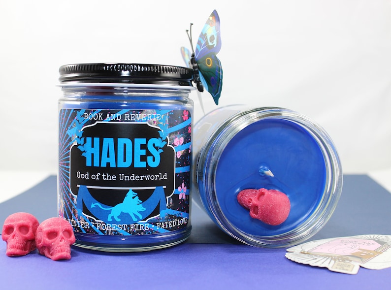 May include: Two glass jar candles with blue wax and a black lid. The label reads "HADES God of the Underworld" with a lion graphic. Pink skull-shaped wax melts and a butterfly decoration are also present.