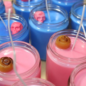 May include: Close-up of several glass jar candles. Some candles are blue with pink skull accents, others are pink with a cinnamon roll accent. Each candle has a white wick. The candles are in clear glass jars.