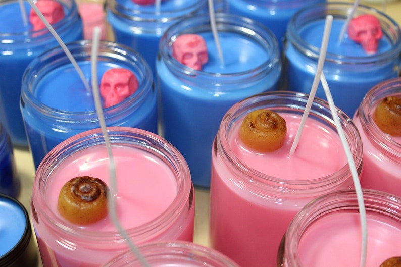 May include: Close-up of several glass jar candles. Some candles are blue with pink skull accents, others are pink with a cinnamon roll accent. Each candle has a white wick. The candles are in clear glass jars.