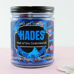 May include: A glass jar candle with a black lid. The label features the word "HADES" in blue, with "God of the Underworld" below. The label also includes the text "BOOK AND REVERIE" and other decorative elements.