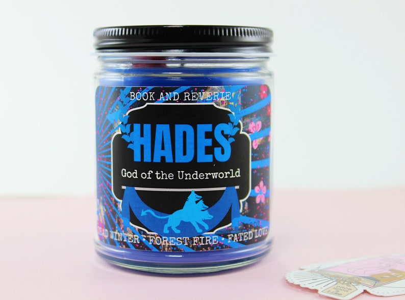 May include: A glass jar candle with a black lid. The label features the word "HADES" in blue, with "God of the Underworld" below. The label also includes the text "BOOK AND REVERIE" and other decorative elements.