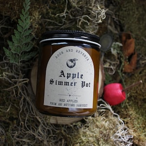 May include: A brown glass jar with a black lid, labeled "Apple Simmer Pot" with the text "Red Apples From An Autumn Harvest." The jar sits on a wooden slice surrounded by moss and a red apple.