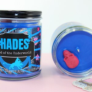 May include: Two glass jar candles with blue wax. One candle is upright, the other is tilted. The label on the upright candle reads "HADES God of the Underworld" with a blue silhouette of a lion. A pink skull is embedded in the wax.