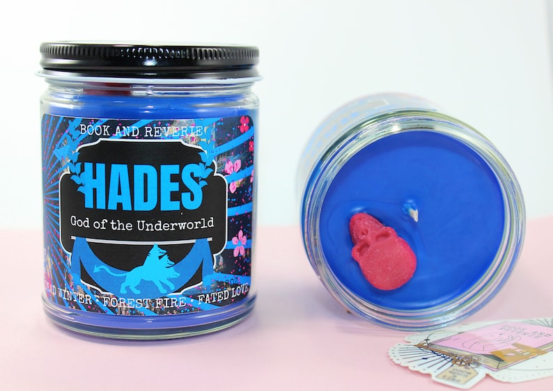 May include: Two glass jar candles with blue wax. One candle is upright, the other is tilted. The label on the upright candle reads "HADES God of the Underworld" with a blue silhouette of a lion. A pink skull is embedded in the wax.