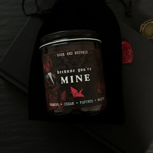 May include: A glass jar candle with a black lid and a label that reads "BOOK AND REVERIE" and "because you're MINE". The label features red roses and a red origami crane. The scent notes are oakmoss, cedar, papyrus, and malfot.
