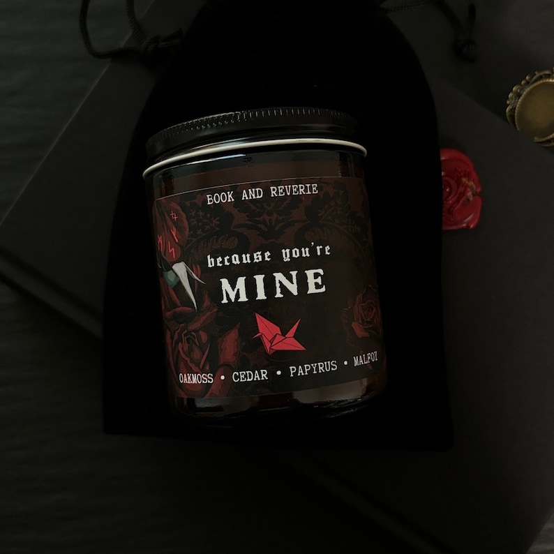 May include: A glass jar candle with a black lid and a label that reads "BOOK AND REVERIE" and "because you're MINE". The label features red roses and a red origami crane. The scent notes are oakmoss, cedar, papyrus, and malfot.
