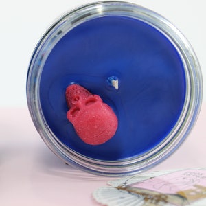 May include: A blue candle in a glass jar with a red skull embedded in the wax. The candle has a wick and is surrounded by a clear glass container. The label on the side of the jar reads "FATED LOVE".