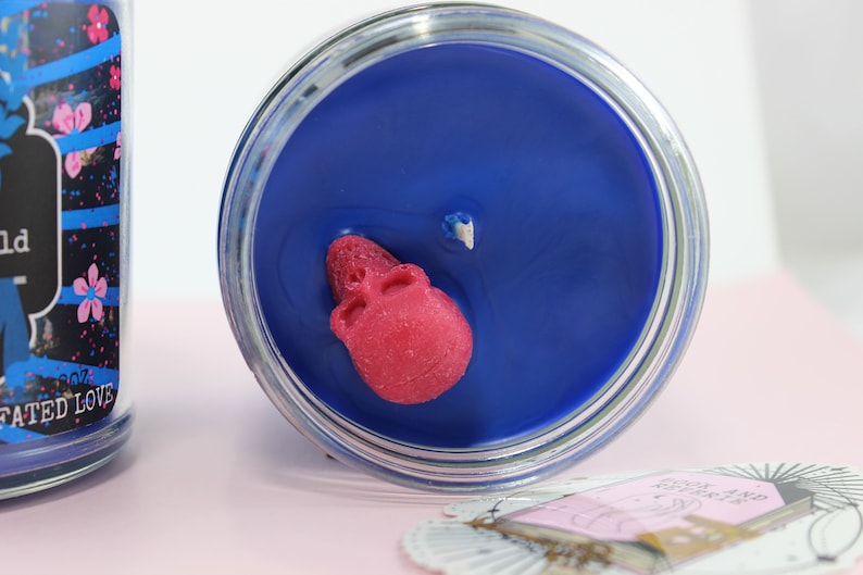 May include: A blue candle in a glass jar with a red skull embedded in the wax. The candle has a wick and is surrounded by a clear glass container. The label on the side of the jar reads "FATED LOVE".