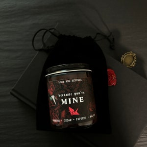 May include: A dark glass jar candle with a black lid, featuring the text "because you're MINE" and the words "BOOK AND REVERIE". The candle is surrounded by a black velvet bag, a black book, and a decorative ring.