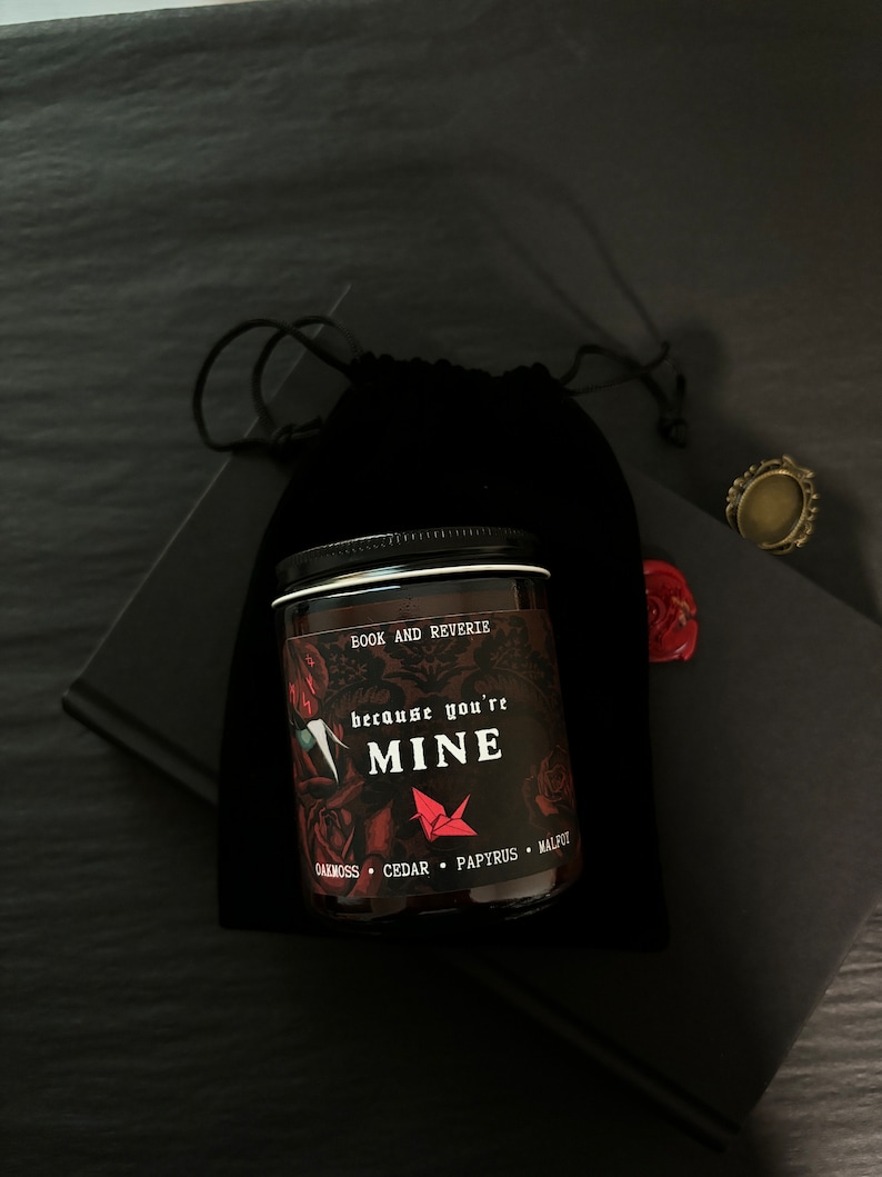 May include: A dark glass jar candle with a black lid, featuring the text "because you're MINE" and the words "BOOK AND REVERIE". The candle is surrounded by a black velvet bag, a black book, and a decorative ring.