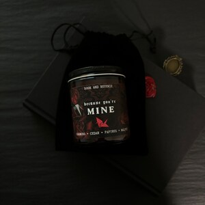 May include: A black candle jar with the text "because you're MINE" and "BOOK AND REVERIE" is placed in front of a black velvet bag. The candle has a black lid and a label with red and white text. A small gold ring and a red wax seal are also visible.