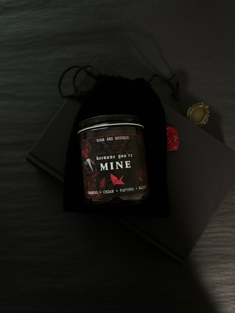 May include: A black candle jar with the text "because you're MINE" and "BOOK AND REVERIE" is placed in front of a black velvet bag. The candle has a black lid and a label with red and white text. A small gold ring and a red wax seal are also visible.