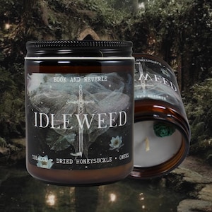May include: Two glass jar candles with black lids. The labels read "IDLEWEED" with the scent notes "TOBACCO, DRIED HONEYSUCKLE, & OAKS". The background is a blurred forest scene. The candle on the right is partially visible, showing the lit wick.