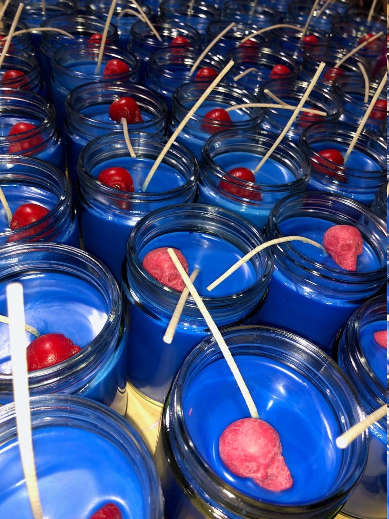 May include: A collection of blue-colored candles in glass jars, each with a red skull-shaped decoration and a white wick. The candles are arranged closely together, creating a visually striking display. The blue wax contrasts with the red skulls.