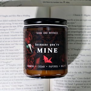 May include: A candle jar with a black lid and a dark label featuring the text "BOOK AND REVERIE" and "because you're MINE." The label includes rose illustrations and the scent notes: Oakmoss, Cedar, Papyrus, and Malfit.