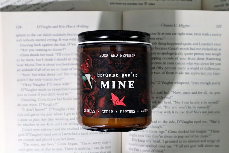 May include: A candle jar with a black lid and a dark label featuring the text "BOOK AND REVERIE" and "because you're MINE." The label includes rose illustrations and the scent notes: Oakmoss, Cedar, Papyrus, and Malfit.