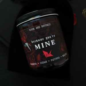 May include: A black jar candle with a black lid, featuring the text "BOOK AND REVERIE" and "because you're MINE" in white. The label includes a red origami crane and rose illustrations. The candle's scent notes are oakmoss, cedar, papyrus, and malpu.