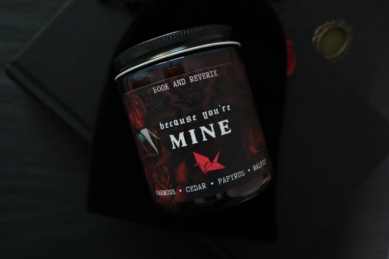 May include: A black jar candle with a black lid, featuring the text "BOOK AND REVERIE" and "because you're MINE" in white. The label includes a red origami crane and rose illustrations. The candle's scent notes are oakmoss, cedar, papyrus, and malpu.