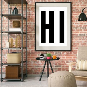 May include: A black framed poster with the word "HI" in large, bold, black letters on a white background. The poster is hanging on a brick wall. There is a small table with a plant and a mug on it, and a brown ottoman in front of the table.