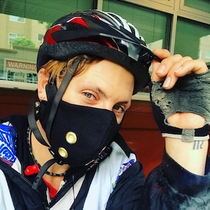 Pode incluir: Uma pessoa usando uma máscara facial preta com botões dourados, um capacete de bicicleta vermelho e preto e uma jaqueta com padrão preto e branco. A pessoa está segurando uma luva preta e cinza com furos.