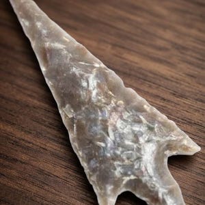 May include: A light gray, translucent flint arrowhead with a sharp, pointed tip and a notched base. The surface features visible knapped edges and a smooth, polished texture with subtle variations in tone throughout the stone material.
