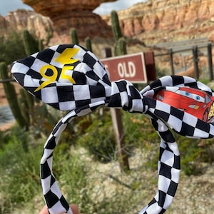 May include: Black and white checkered headband with a yellow lightning bolt and a red cartoon car on one side. The headband is tied in a bow.