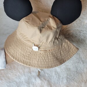 Puede incluir: Un sombrero de sol de color beige con orejas de Mickey Mouse negras. El sombrero tiene un bot&oacute;n blanco y un cord&oacute;n.