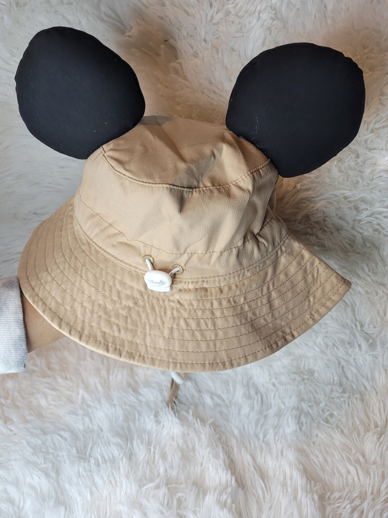 Puede incluir: Un sombrero de sol de color beige con orejas de Mickey Mouse negras. El sombrero tiene un bot&oacute;n blanco y un cord&oacute;n.