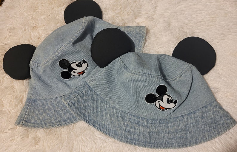 May include: Two denim bucket hats with black Mickey Mouse ears and embroidered Mickey Mouse faces. The hats are light blue and have a white background for the Mickey Mouse faces.
