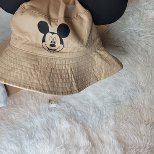 Puede incluir: Un sombrero de cubo de color beige con orejas de Mickey Mouse negras y un gr&aacute;fico de la cara de Mickey Mouse negro en la parte delantera.