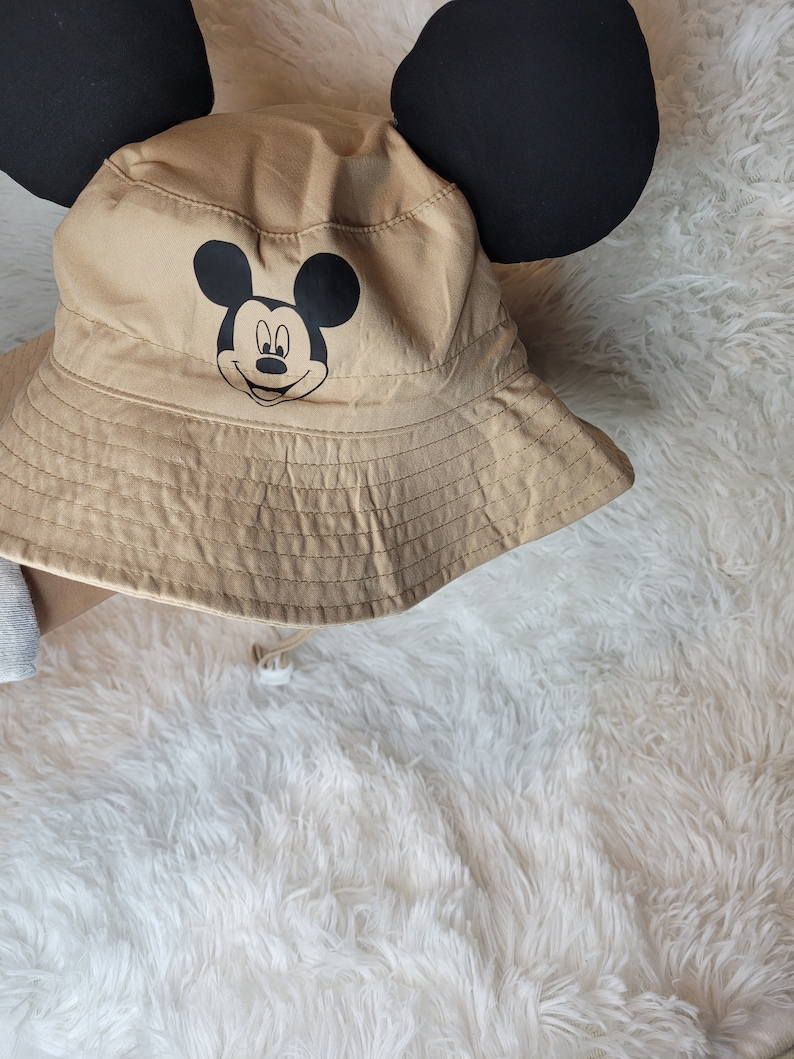 Puede incluir: Un sombrero de cubo de color beige con orejas de Mickey Mouse negras y un gr&aacute;fico de la cara de Mickey Mouse negro en la parte delantera.