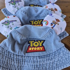 May include: Several light blue denim bucket hats with the "Toy Story" logo embroidered on the front. Each hat has attached ear-shaped accents with colorful cartoon character designs, including Buzz Lightyear and Woody.