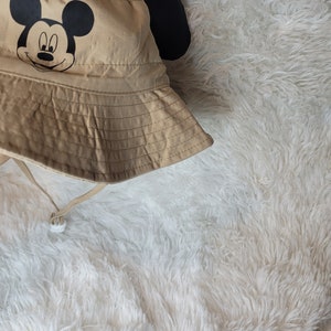 Puede incluir: Un sombrero de cubo de color beige con orejas de Mickey Mouse negras y una cara de Mickey Mouse negra en la parte delantera.