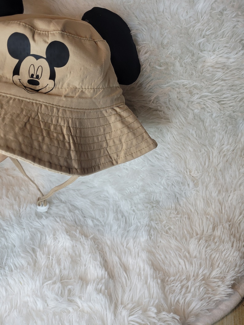 Puede incluir: Un sombrero de cubo de color beige con orejas de Mickey Mouse negras y una cara de Mickey Mouse negra en la parte delantera.