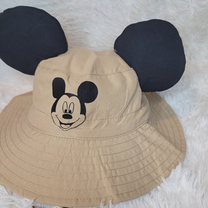 Puede incluir: Un sombrero de cubo de color beige con orejas de Mickey Mouse negras y una cara de Mickey Mouse negra en la parte delantera.