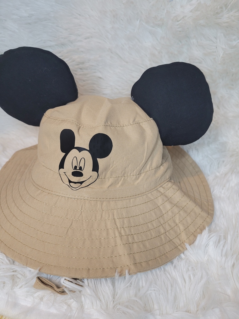 Puede incluir: Un sombrero de cubo de color beige con orejas de Mickey Mouse negras y una cara de Mickey Mouse negra en la parte delantera.