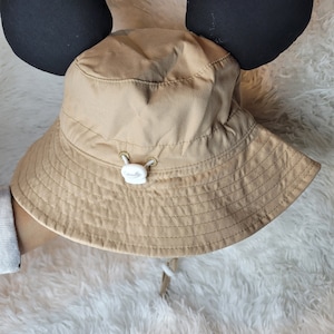 Puede incluir: Un sombrero de cubo de color beige con orejas de Mickey Mouse negras adjuntas. El sombrero tiene un bot&oacute;n blanco y un cord&oacute;n blanco.