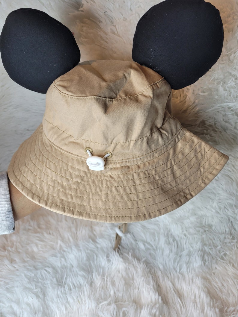 Puede incluir: Un sombrero de cubo de color beige con orejas de Mickey Mouse negras adjuntas. El sombrero tiene un bot&oacute;n blanco y un cord&oacute;n blanco.