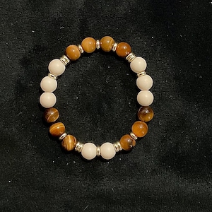 May include: A beaded bracelet featuring alternating light beige and tiger's eye beads. The bracelet is strung with silver-colored spacers. The beads are round and have a polished finish. The bracelet is displayed on a dark background.