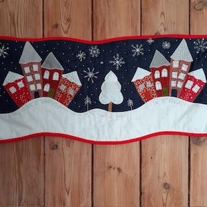 May include: A winter-themed wall hanging with a navy blue background, featuring red houses with gray roofs and white snow. The phrase "Let it snow..." is written in cursive. The piece is trimmed with a red border and has a white snow-covered base.